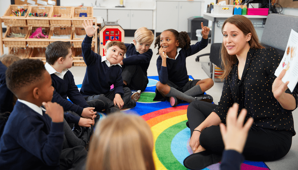 Primary school children sitting on the floor in a classroom with their teacher, raising hands to answer a question.