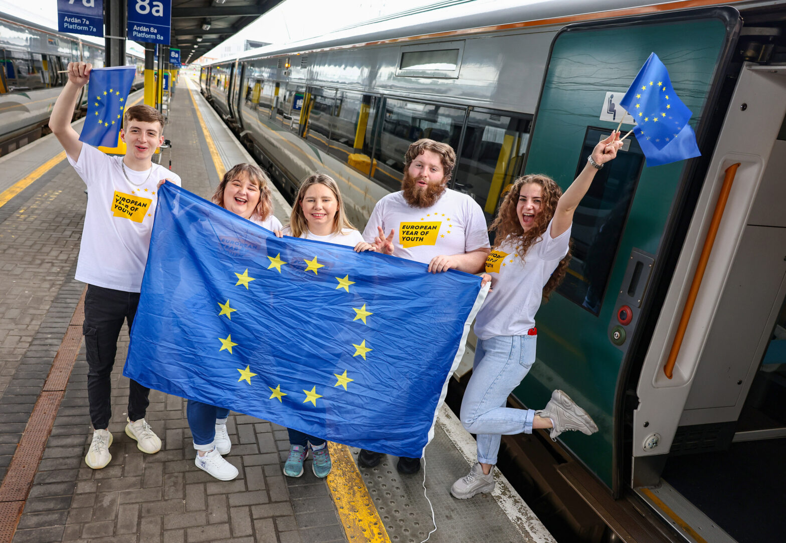 Promotional image for European Year of Youth. A group of young people on a train platform holding a European flag and wearing t-shirts that read european year of youth