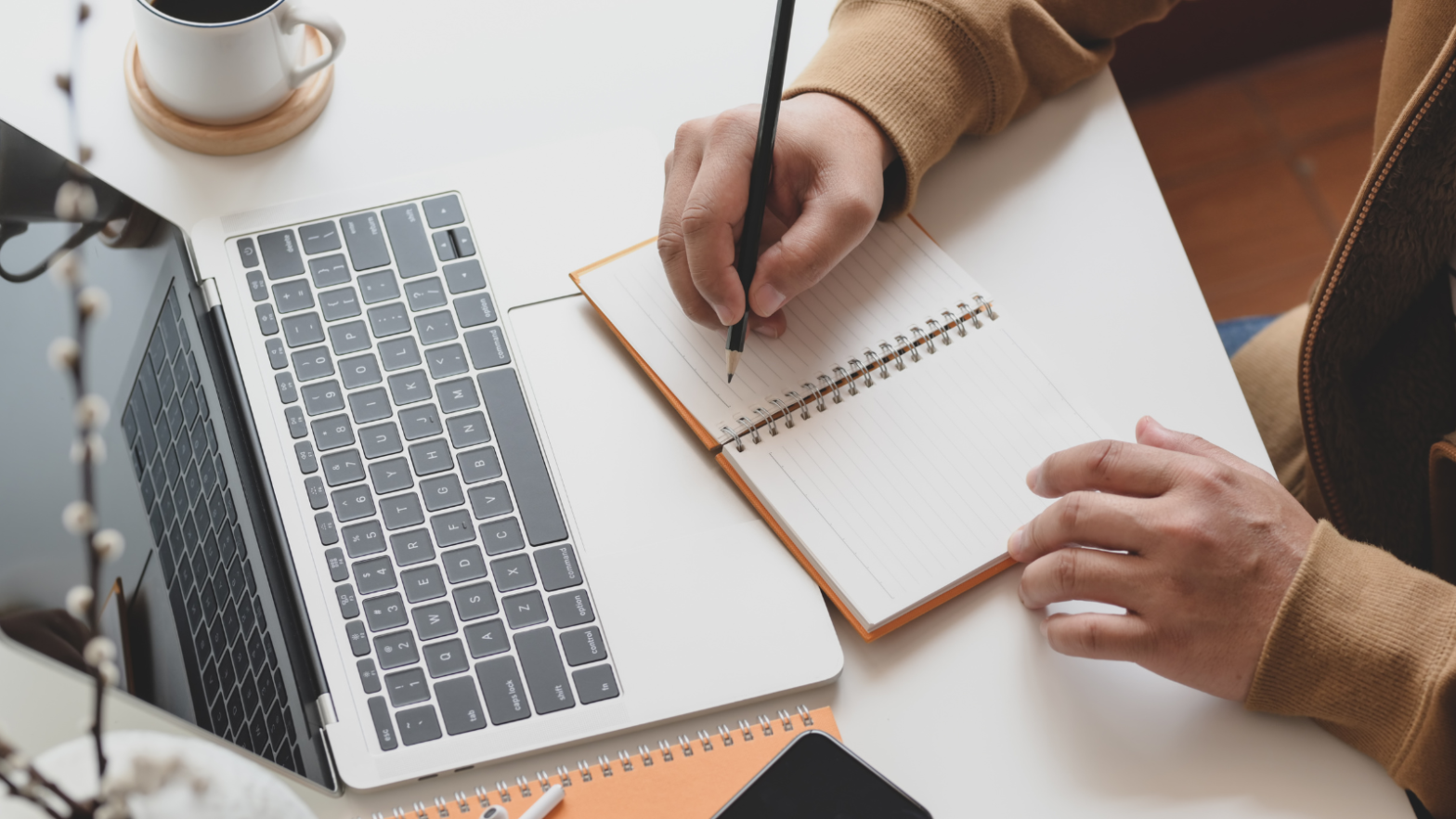 A stock image of a desk with an open laptop and a student writing on his notebook